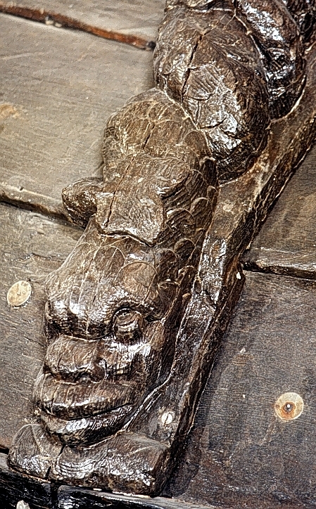 Triton med spetsigt, krusigt skägg och stora mustascher. På huvudet sitter en hjälm med stora plymer och vaga spår av en djurmaskaron. På hjälmens framsida sitter en rullverksvolut. Överkroppens övergång till fiskstjärten markeras av två stora, dekorativa bladslingor med upprullade ändpartier. Bladslingorna är lagda över varandra så att de bildar något som närmast kan liknas vid ett vävt mönster. Skulpturens fiskstjärt slingrar sig kring stjärten till en fisk eller delfin, vars huvud är synligt vid skulpturens fotända.

Bakstycket är kraftigt och snidat i ett med skulpturen. Baksidan är försedd med hak eller urtag. Skulpturens profil är svagt S-formad.
Skulpturen är tämligen välbevarad.

Text in English: Triton with pointed curly beard and large moustache. The sculpture wears a helmet with a large plume and vague traces of an animal mask and scroll volutes on the front side. The transition from the lower body to the fish tail is marked by two large, decorative leafy vines, the ends of which are rolled up and laid across each other so as to form an almost woven pattern. The fish tail is entwined with the tail of a fish or dolphin whose head can be seen at the foot of the sculpture.

The backboard is robust and carved in one piece with the figure. The back side of the backboard was carved with hollowed out grooves. The profile is slightly S-shaped.
The sculpture is rather well preserved.