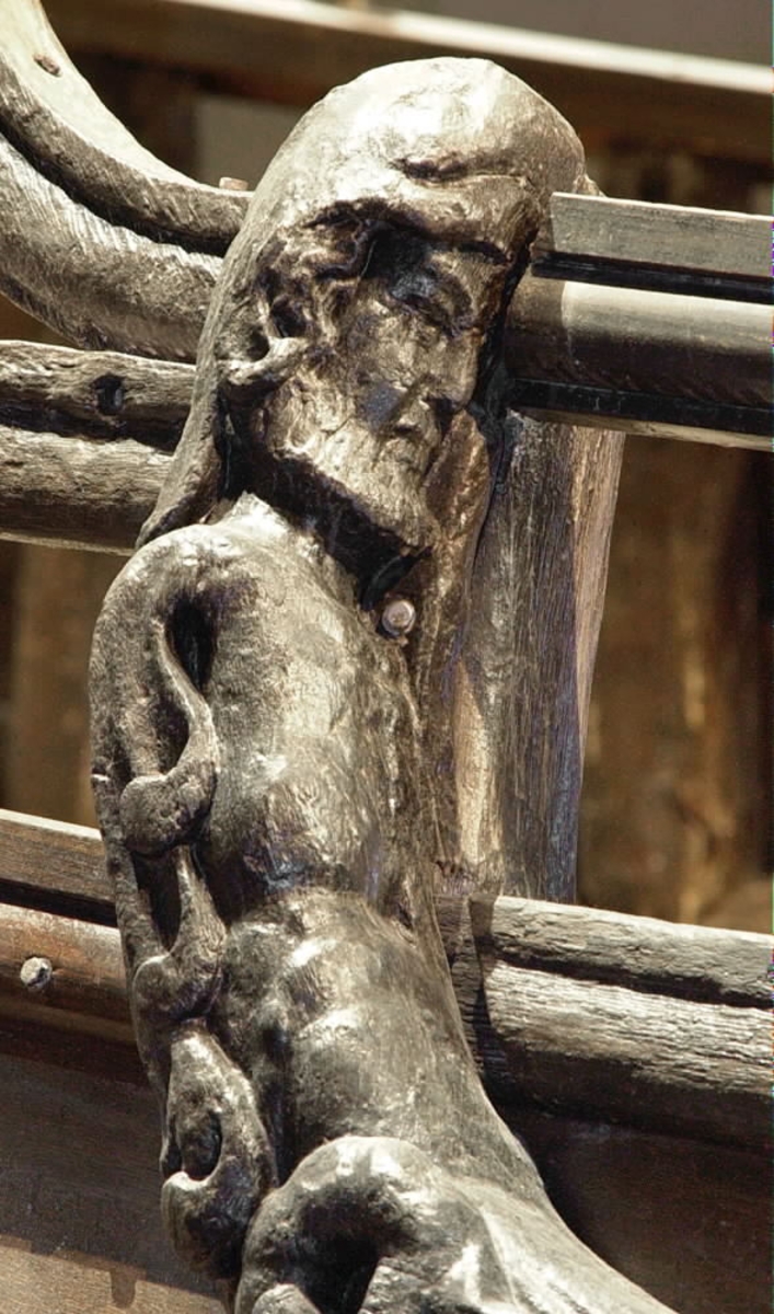 Skulptur föreställande en triton, svängd mot vänster och framställd i höger profil.
Tritonen har skäggigt ansikte, stor näsa samt en bladsmyckad hjälm på huvudet. Armarna är ersatta av långa, flikiga bladslingor. Bålens övergång i den fjällförsedda fiskstjärten markeras av stora bladbildningar. Figuren avtecknar sig i hög relief mot det släta bakstycket. Nedtill ses en fiskliknande stjärt med drakfena, vilken är fortsättningen på en under liggande draken, se fyndnummer 00628. På baksidan finns urtag för relingslisten.
Skulpturen är välbevarad.

Text in English: A sculpture of a Triton curved to the left, in right profile.
Bearded face with a long nose. Leaf-decorated helmet. The arms are replaced by long, lobed leaf formations. The transition between the male torso and the scaled fish-tail is decorated with large leaves. The figure is seen in high relief against a plain back board. Below the figure can be seen a section of another fish-like tail but actually a dragon''s fin, belonging to the dragon situated underneath, see No. 00628. There are notches on the back of the sculpture that fit into the railing.
The sculpture is well preserved.