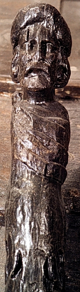 Skulpterad Triton med tjock lagerkrans på huvudet.
Figuren har stora yviga mustascher och långt, lockigt hår. Snett över bröstet är en klädnad draperad. Klädnaden är hopfäst på vänster axel.

Övergången till de snodda fiskstjärtarna döljs av ett stort, flikigt blad vars mittflik är förtjockad och upprullad. Skulpturens fotända är tapp- eller kilformad och tillplattad från sidorna. Tritonens huvud är framställt som rundskulptur. Från axlarna och nedåt är baksidan slät och plan. I höjd med övergången till stjärtpartiet finns ett kraftigt urtag. Nedtill bryter baksidans fotända av inåt.

Text in English: Sculpted Triton with thick laurel wreath on its head.
The figure has a large bushy moustache and long curly hair. A garment is draped across the chest and fastened on the left shoulder.

The transition between the upper body and fish tails is concealed by a large leaf, the middle segment of which is thickened and rolled up. The foot end of the sculpture has a flat end, seen from the side, and is wedge-shaped. The Triton''s head is carved as a three-dimensional sculpture. The rear side is smooth. There is a deep groove on the rear side at the transistion point between the male torso and the fish tails.