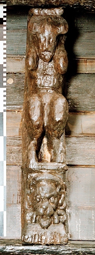 Skulptur föreställande en djävulsfigur i starkt hukande ställning, placerad på en hög sockel.
Figurens huvud är utrustat med stora, krumma horn. I vardera handen håller den på ömse sidor av huvudet något som möjligen kan tolkas som en stor snäcka eller lur. Hakan pryds av ett bockskägg. Figuren har fladdermusvingar på ryggen och starkt luden underkropp med klövförsedda bockfötter. Fötterna vilar på den starkt sluttande sockeln, som framtill är prydd med en ap- eller lejonliknande maskaron med stor nos och hög, välvd panna. På sockelns sidor ses voluter.
Sockeln utgör fortsättning på det kraftiga klink-konstruerade bakstycket som är framdraget ovanför figuren.
Skulpturen är delvis kraftigt sliten och vittrad, särskilt den övre delen. Nederdelen är något bättre bevarad.

Text in English: Sculpture of a devil figure in a crouched position, placed on a high socle.
The figure''s head is equipped with large, curved horns. In either hand the figure holds on either side of its head something that may possibly be identified as a large seashell or horn. The devil has a goat''s beard on its chin, bat wings on its back and very hirsute lower parts of the body, and cloven goats'' feet.
The feet rests on a sharply sloping socle that is decked in front with a mask of an ape-like or lion-like face with a large nose and a high arched forehead. On the sides of the socle volutes are visible.
The socle forms the extension of the heavy, clinker-built back-piece, which is drawn forward above the figure.
The sculpture, particularly its upper parts, is much worn and disintegrated. The lower part is somewhat better preserved.