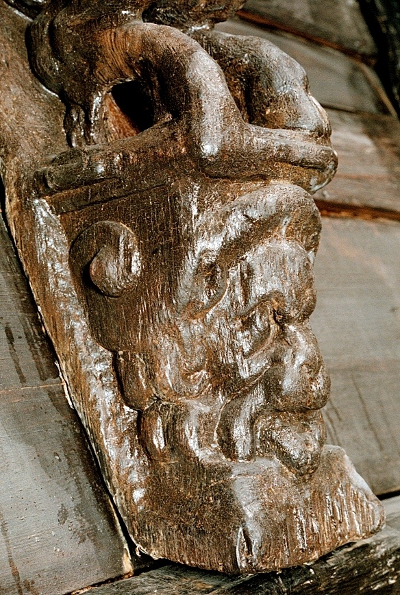 Skulptur föreställande en djävulsfigur i starkt hukande ställning, placerad på en hög sockel.
Figurens huvud är utrustat med stora, krumma horn. I vardera handen håller den på ömse sidor av huvudet något som möjligen kan tolkas som en stor snäcka eller lur. Hakan pryds av ett bockskägg. Figuren har fladdermusvingar på ryggen och starkt luden underkropp med klövförsedda bockfötter. Fötterna vilar på den starkt sluttande sockeln, som framtill är prydd med en ap- eller lejonliknande maskaron med stor nos och hög, välvd panna. På sockelns sidor ses voluter.
Sockeln utgör fortsättning på det kraftiga klink-konstruerade bakstycket som är framdraget ovanför figuren.
Skulpturen är delvis kraftigt sliten och vittrad, särskilt den övre delen. Nederdelen är något bättre bevarad.

Text in English: Sculpture of a devil figure in a crouched position, placed on a high socle.
The figure''s head is equipped with large, curved horns. In either hand the figure holds on either side of its head something that may possibly be identified as a large seashell or horn. The devil has a goat''s beard on its chin, bat wings on its back and very hirsute lower parts of the body, and cloven goats'' feet.
The feet rests on a sharply sloping socle that is decked in front with a mask of an ape-like or lion-like face with a large nose and a high arched forehead. On the sides of the socle volutes are visible.
The socle forms the extension of the heavy, clinker-built back-piece, which is drawn forward above the figure.
The sculpture, particularly its upper parts, is much worn and disintegrated. The lower part is somewhat better preserved.