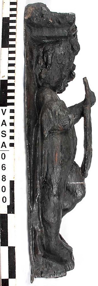 Skulptur föreställande en barnfigur spelande på en viola da braccio.
Fiolen hålles med vänster hand och stöder mot vänster knä. Kroppstyngden vilar på höger ben. Huvudet är vridet åt höger. Antydan till mantel utmed höger sida. Ovanför hjässan ses en äggstavslist. Figuren står på ett minimalt underlag som troligen avser att föreställa ett stycke naturlig mark. Skulpturens baksida är slät
Skulpturen är kraftigt sliten. Höger underarm är bortbruten.

Text in English: This is a sculpture of a child holding a viola da braccio in the left hand and supported on the left knee.
The weight of the body rests on the right leg. The head is turned to the right. There is the suggestion of a mantle on the figures right side. Just above the head, the back board comes forward at right angles and the front of the board is decorated with an egg moulding design. The figure stands upon a small plinth. The back of the sculpture is smooth.
The sculpture is very worn and the right forearm is broken off.