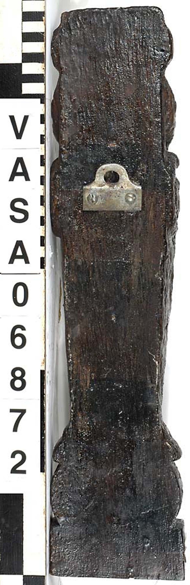 Liten skulpterad pilaster, troligtvis utformad som en karyatid eller möjligen en putto. Figuren har långt hår och ett knubbigt ansikte, omgivet av en bred krage. Det nedåt avsmalnande pilasterskaftet är dekorerat med akantusblad. Nedtill avslutas pilastern av en rikt profilerad och dekorerad sockel. Skulpturens baksida är slät.

Skulpturen är sliten, pilasterns nedre del är dock mindre sliten än den övre.



Text in English: A small carved pilaster, with a caryatid or probably a putto. The figure has a chubby face, long hair and a broad collar. The lower part of the pilaster is decorated with acanthus leaves. The caryatid stands on a richly profiled and decorated socle. The back of the sculpture is smooth.

The sculpture is worn, the lower end less so than the upper.