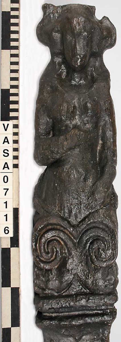 Skulpterad pilaster med övre delen i form av en karyatid med yppig, konstrik frisyr.
Höger arm hålls böjd med handen placerad framför bröstet. Vänster arm hålls utsträckt utefter sidan med handen vilande mot höften. Underkroppen döljs av ett hölje som framtill är prytt med stora, spiralformade ornament och små djurfigurer.
Karyatiden avgränsas från det rullverksornerade pilasterskaftet av en profilerad list. Nedtill avslutas skaftet av ett rullverksornament ovanför den rikt profilerade basen. Nedtill är pilastern försedd med en hög piedestal, prydd med beslagsornamentik. Figuren och dekoren omfattar även pilasterns sidor. Baksidan är slät.


Text in English: Sculpted pilaster with its upper part shaped as a Caryatid with a luxuriant artistic coiffure.
The right arm is held bent with the hand in front of the breast, the left arm is held at the side with the hand resting against the thigh. The lower part of the body is concealed by a shift or slip that is decorated with large spiral-shaped ornaments and small figures of animals.
A profiled batten separates the Caryatid from the shaft of the pilaster, which is ornamented with scrollwork. The shaft terminates with a scrollwork ornament above the richly profiled base. Below, the pilster is equipped with a high pedestal, decked with applied ornamentation. The back is smooth.