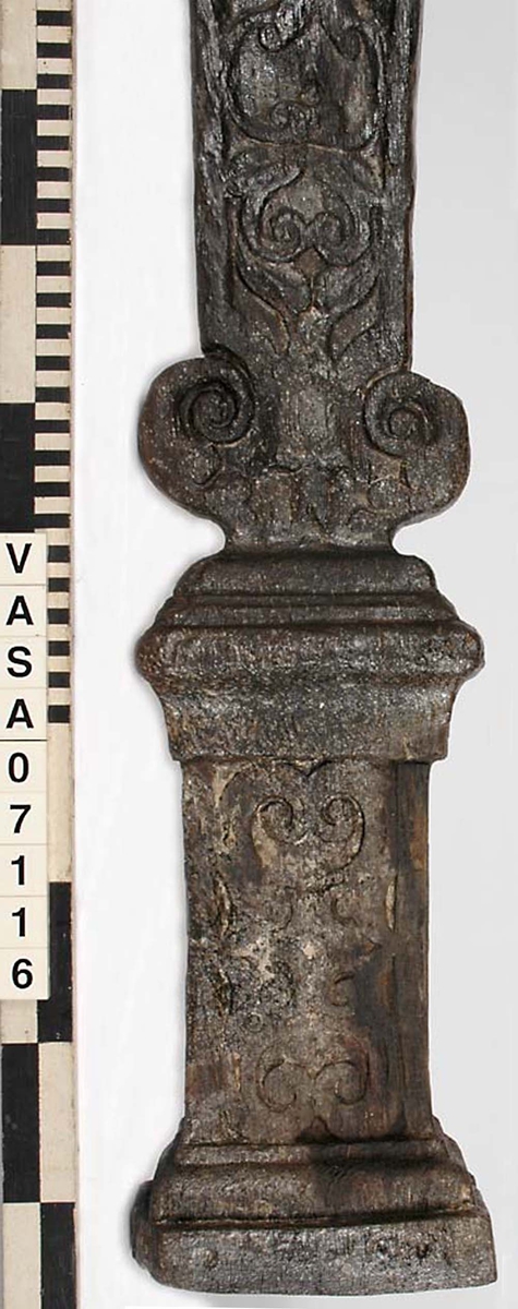 Skulpterad pilaster med övre delen i form av en karyatid med yppig, konstrik frisyr.
Höger arm hålls böjd med handen placerad framför bröstet. Vänster arm hålls utsträckt utefter sidan med handen vilande mot höften. Underkroppen döljs av ett hölje som framtill är prytt med stora, spiralformade ornament och små djurfigurer.
Karyatiden avgränsas från det rullverksornerade pilasterskaftet av en profilerad list. Nedtill avslutas skaftet av ett rullverksornament ovanför den rikt profilerade basen. Nedtill är pilastern försedd med en hög piedestal, prydd med beslagsornamentik. Figuren och dekoren omfattar även pilasterns sidor. Baksidan är slät.


Text in English: Sculpted pilaster with its upper part shaped as a Caryatid with a luxuriant artistic coiffure.
The right arm is held bent with the hand in front of the breast, the left arm is held at the side with the hand resting against the thigh. The lower part of the body is concealed by a shift or slip that is decorated with large spiral-shaped ornaments and small figures of animals.
A profiled batten separates the Caryatid from the shaft of the pilaster, which is ornamented with scrollwork. The shaft terminates with a scrollwork ornament above the richly profiled base. Below, the pilster is equipped with a high pedestal, decked with applied ornamentation. The back is smooth.