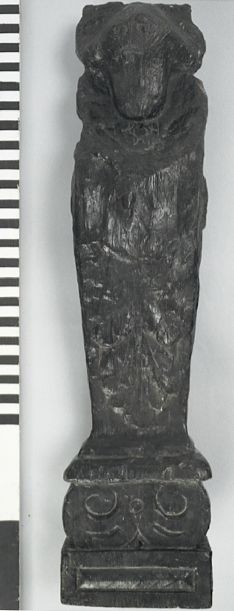 Liten skulpterad pilaster, utformad som en karyatid eller möjligen en putto. Figuren har långt hår och ett knubbigt ansikte, omgivet av en bred krage. Det nedåt avsmalnande pilasterskaftet är dekorerat med akantusblad. Nedtill avslutas pilastern av en rikt profilerad och dekorerad sockel. Skulpturens baksida är slät.
Skulpturen är sliten, främst den övre delen.

Text in English: A small carved pilaster, with a caryatid or probably a putto. The figure has a chubby face, long hair and a broad collar. The lower part of the pilaster is decorated with acanthus leaves. The caryatid stands on a richly profiled and decorated socle. The back of the sculpture is smooth.
The sculpture is worn.