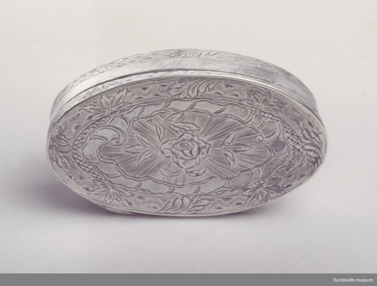 Snusdosa i silver. Oval form, invändigt förgylld. På locket och runt sidorna graverade och svisselerade brodyrer med blommor och blad. En graverad ros på locket. På framsidan är 'M:Wld' graverat.