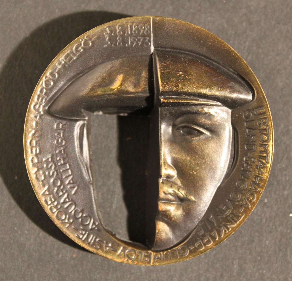Medalj i två delar av patinerad brons. Åtsidans
yttersidavisar ett halvporträtt av kung Gustaf VI Adolf. Till vänster
synskungens hand hålla en urna, med text enligt MRK. Åtsidans
insidavisar den unge prins Gustaf Adolfs halvporträtt. Texten upptar
ettantal platser där utgrävningar skett i närvaro av eller på
initiativfrån kungen. Frånsidans yttersida visar nederst kungen inför
enutgrävning. Över hela motivet framtonar en kvinnokropp
symboliserandeModer Jord. Frånsidans insida visar ett tvådelat
ansiktsporträtt avden unge prinsen i nedsänkt relief till vänster och
Gustaf VI Adolf iupphöjd relief till höger, med text "Svenska
Arkeologiska SamfundetsHöge Beskyddare". Medaljen firar kungens 75 år
som arkeolog. Den gavsut av Svenska Arkeologiska Samfundet 1973. På
randen står medaljenslöpande tillverkningsnummer och tillverkaren
Sporrong instansat.