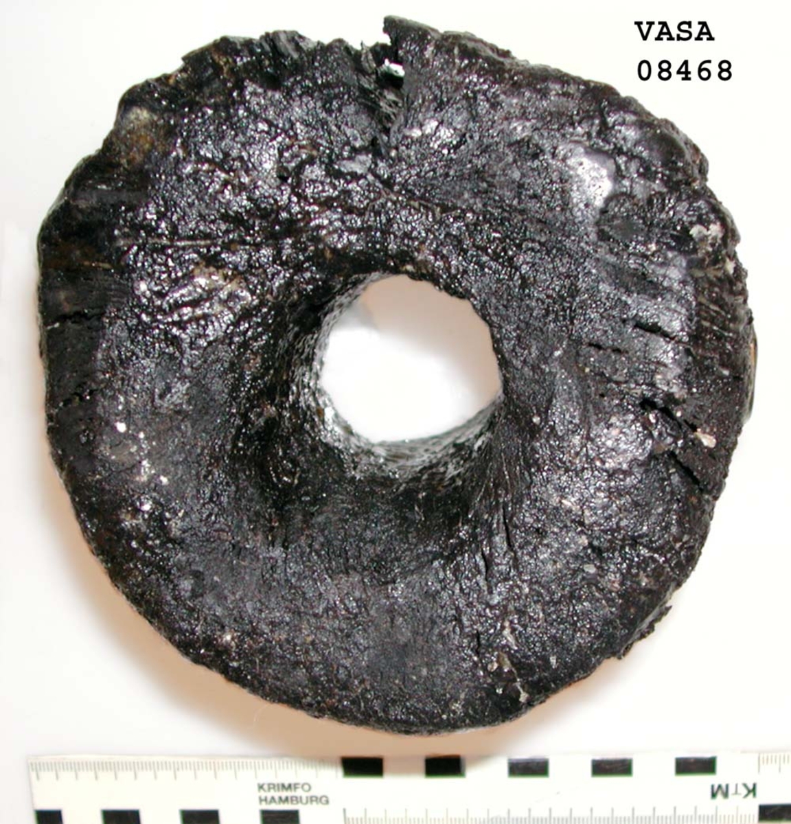 Kolv till ansättare. Kolven består av ett cylindriskt svarvat trästycke med ett centrumhål för skaftet. I kolvens övre ände, mot skaftet till, är kolven profilsvarvad. Kolven är kraftigt eroderad och sprucken. Profilsvarvningen upptill är nedsliten.