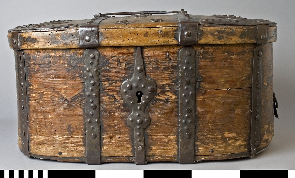 Färdskrin av trä med beslag av järn. Oval form med ett lock som är dekorerat med smida järnbeslag. Kanten på locket är skod med järn. På framsidan löper beslag ned och runt om. Centrerat mitt fram finns ett lås med nyckelskylt i järn.