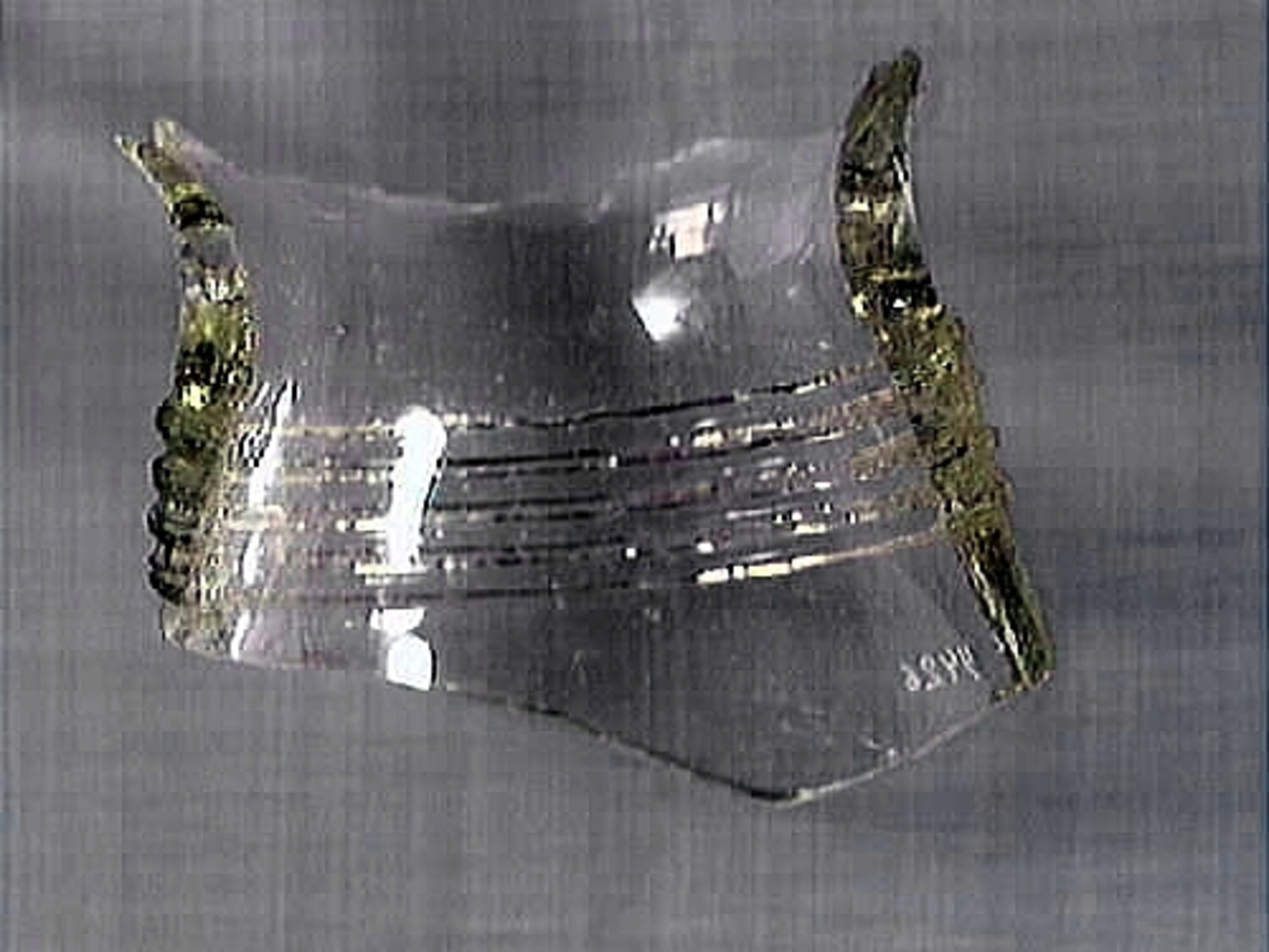 En glasskärva. Troligtvis härrör skärvan från ett dricksglas, eventuellt en flaska. Skärvan är försedd med en banddekor som består av tre glassträngar. Glaset är transparant och svagt grönfärgat.

2015: No damage. Glass is clear with a greenish.  Glass has three bands running horizontally.