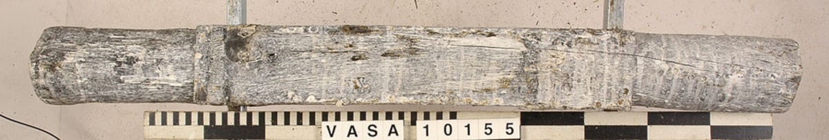 Lavettaxel, främre. Ett avlångt trästycke med kvadratiskt tvärsnitt mitt på samt cylinderformade ändar. Axeln var fastsatt i lavettbottnens undersida med två grova spikar och två järnband.

För mer information se Dokumentationsblankett under Referenser – Länkade filer.

Text in English: Axle (front) for gun carriage 10153, consisting of a square-section beam with round stub axles at the ends, originally fixed to a notch in the underside of the carriage bed by two nails and two iron straps.

For more information see the Documentation form under References – Linked files.