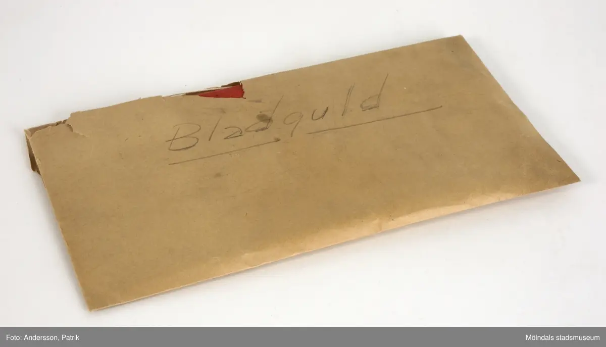 Bladguld i silkespapper, förvaras i pappfodral. 1: Röd pappärm (160x117 mm), textilklädd. Innehåller två små silkespappershäften, vari bladguldet ligger.2: Naturvitt papperkuvert (105x180 mm), som innehåller två små silkespappershäften, vari bladguldet ligger.3: De ovannämnda föremålen förvaras i ett avlångt, brunt kuvert, märkt "Bladguld" med blyerts. Kuvertet är mycket skört. 