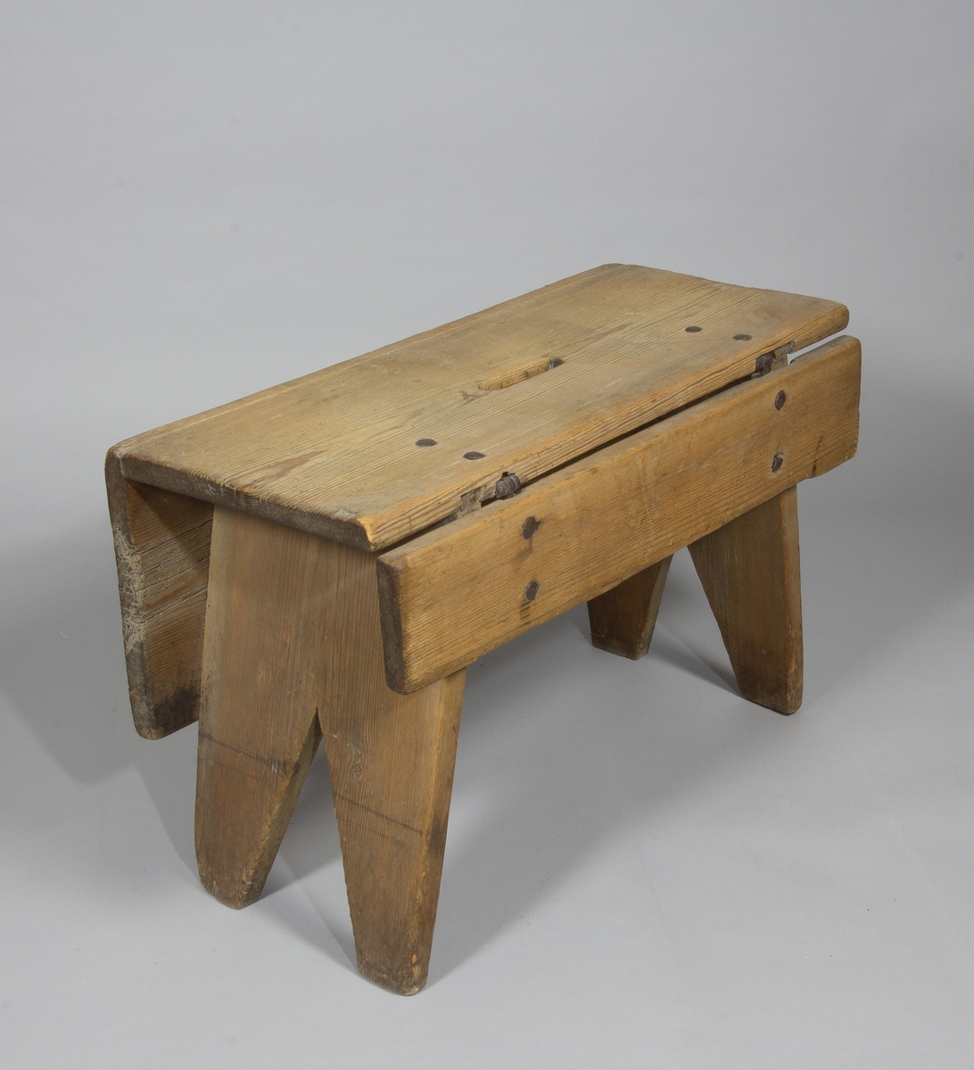 Pall eller pallbol i furu. Låg, avlång med bockformade ben. På pallens ena långsida väl tilltagen sarg, på andra sidan av fälltyp, fäst med gångjärn. Handtag i sätet i form av ett hål.