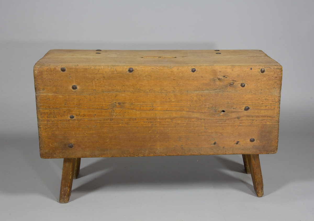 Pall eller pallbol i furu. Låg, avlång med bockformade ben. På pallens ena långsida väl tilltagen sarg, på andra sidan av fälltyp, fäst med gångjärn. Handtag i sätet i form av ett hål.