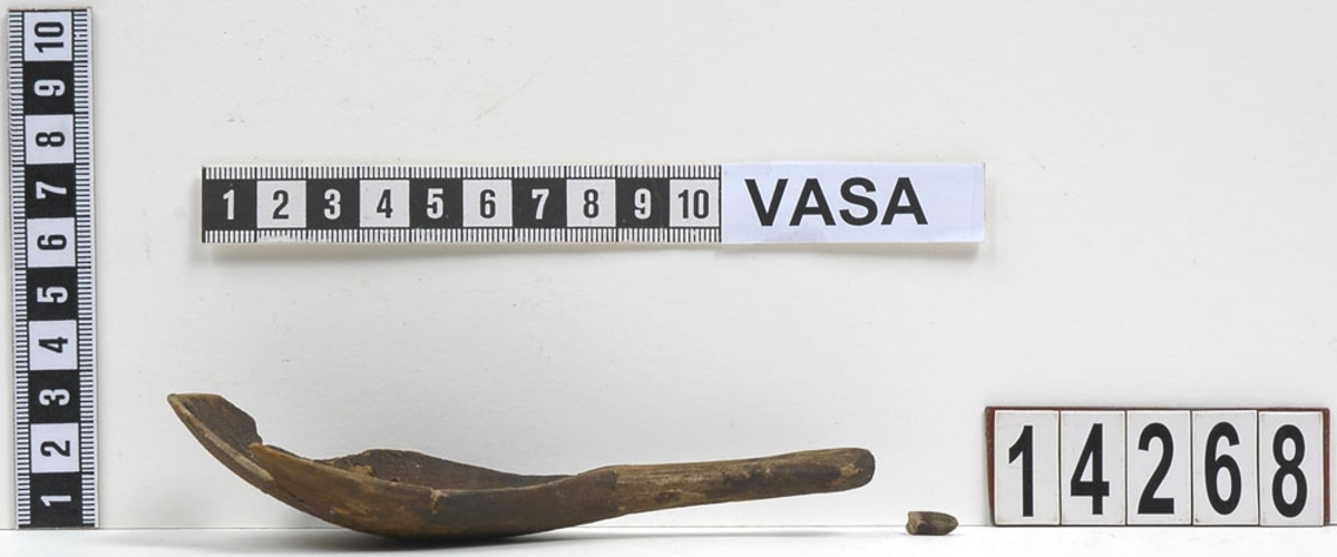 En träsked, ursprungligen bruten i sex delar. Den största delen utgjordes av skedskaftet samt en del av skedbladet. Fyra av delarna tillhörde skedbladet. Den minsta delen utgjordes av en liten knopp, vilken suttit i skedskaftets ände. Skeden är idag sammanfogad av konserveringsenheten.
Skedbladet är äggformat och relativt stort i jämförelse med skaftet. På insida av skedbladet - längs med ytterkanterna, intill skaftet - syns en inristad dekor i form av linjer och streck. Skaftet är kort och rakt, med profilerad ände. På skedbladets baksida, intill skaftet, är ett bomärke inristat. På skeden syns rikliga spår av rödbrun färg.
Skeden är mycket ömtålig. Skedbladets ytterkanter saknas delvis.