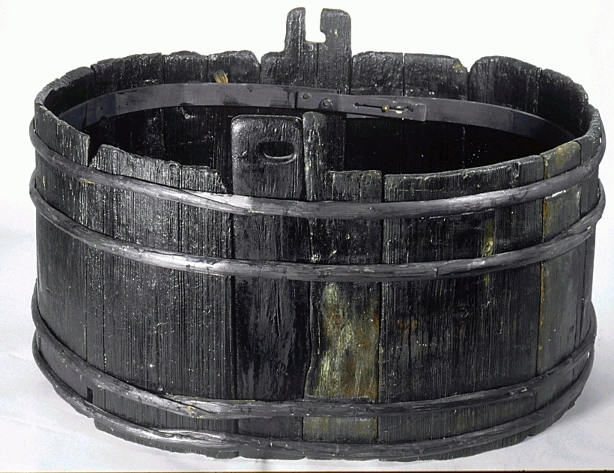 Laggstav till en balja. I stavens övre del finns ett urtag för en regel eller tvärslå. Urtaget är skadat och den övre delen saknas. Runt urtaget finns en fasad nedsänkning, eventuellt för någon form av beslag. På insidan av stavens nedre ände finns ett urtag. Urtaget är ett hugget spår där baljans bottenplatta sitter.