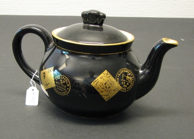 Tekanna i svart porslin med frimärksmotiv och stämpelavtryck
i guldfärg:  Nya Sverige Minnet 5 öre, 2 x 60 öre och 20 öre, 1938,
stämpelavtryck med text: Stockholm Nya Sverige minnet Stadshuset;
Sjöpostexp 7 Nya Sverige minnet.