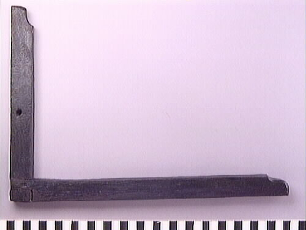 En vinkelhake.

Vinkelhaken består av två plana stavar som är sammanfogade genom sinkning. Ändarna är profilerade. I mitten av den kortare staven finns ett runt hål för upphängning.

Vinkelhaken är välbevarad och i gott skick.

För mer information se Dokumentationsblankett under Referenser – Länkade filer.