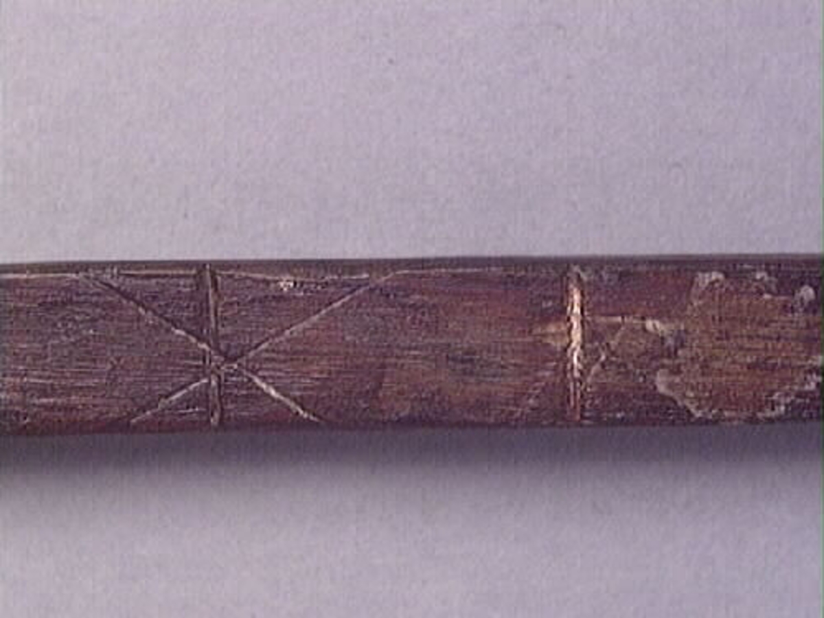 En tumstock av trä.

Tumstocken är försedd med elva inristade skåror som markerar tolv tum. På den mittersta skåran finns ett inristat kryss. Ytterligare ett kryss, dock något mindre, har ristats in vid skåran närmast mittskåran.

Tumstocken är i gott skick.

För mer information se Dokumentationsblankett under Referenser – Länkade filer.