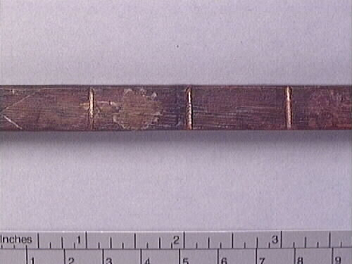 En tumstock av trä.

Tumstocken är försedd med elva inristade skåror som markerar tolv tum. På den mittersta skåran finns ett inristat kryss. Ytterligare ett kryss, dock något mindre, har ristats in vid skåran närmast mittskåran.

Tumstocken är i gott skick.

För mer information se Dokumentationsblankett under Referenser – Länkade filer.