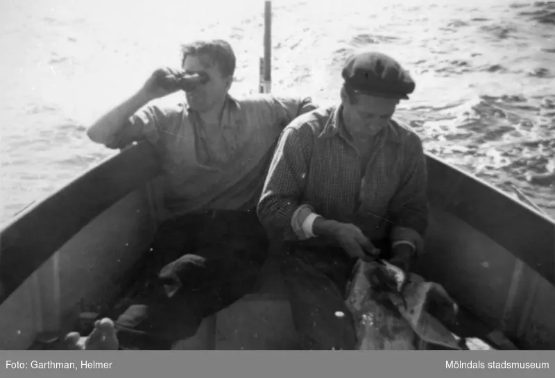 Helmer Garthmans arbetskompisar är ute med båt och fiskar. En man tittar i kikare utöver vattnet och den andre rensar fisk på ett tidningspapper. Näset, 1950-tal. Skänkt av sonen Alf Garthman som arbetade under många år på Mölndals stadsmuseum.