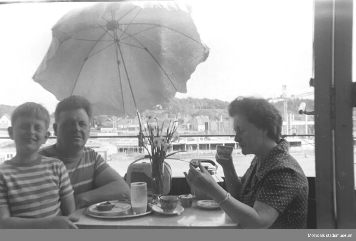 Familjen Garthman sitter på balkongen och fikar. Från vänster Jerry, Helmer och Astrid på Lerbrännargatan 1F, 1963. I bakgrunden skymtar lyftkranar och nybygget av Idrottshuset (senare Aktiviteten). Bygget pågick 1962-1965.