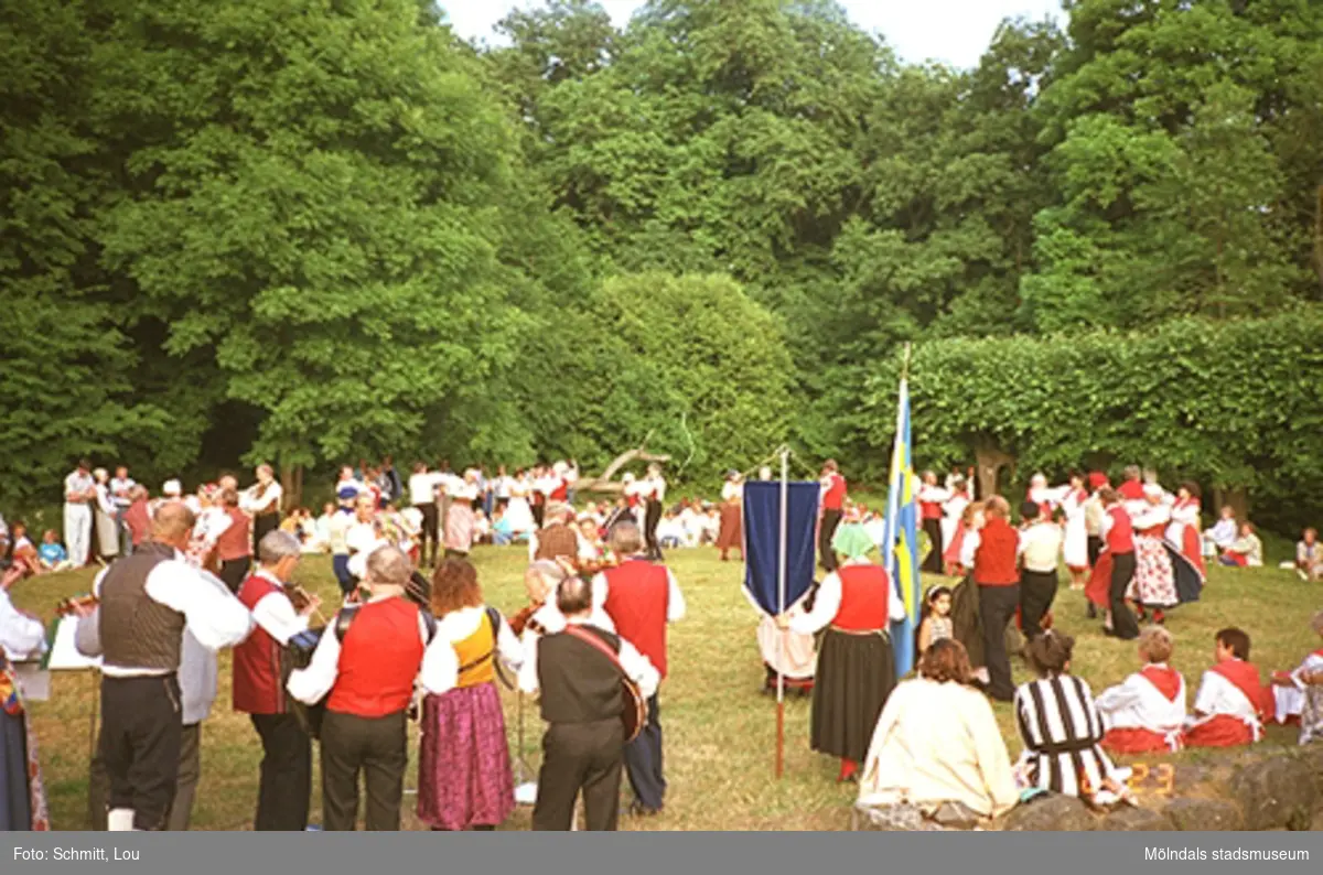 Ett folkdanslag, uppklädda i dräkter, dansar inför beskådare. En kvinna står och håller en blå vimpel i ena handen och en fana, av Svenska flaggan, i andra handen. Gunnebo slottspark 1988.