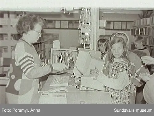 Dokumentationsfotografering av verksamheten vid biblioteksfilialen i Matfors. Bild 01Biblioteksassistenten Ingrid Engström, Matfors bibliotek, med låntagare vid utlånedisken. Bild 09 bibliotekaren Inger Barkman läser för en grupp förskolebarn i bibliotekets "läsrum".