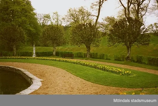 Till vänster ses en del av dammen. Till höger ses parken med träd, blomsterrabatt, en skulptur och grusgångar.