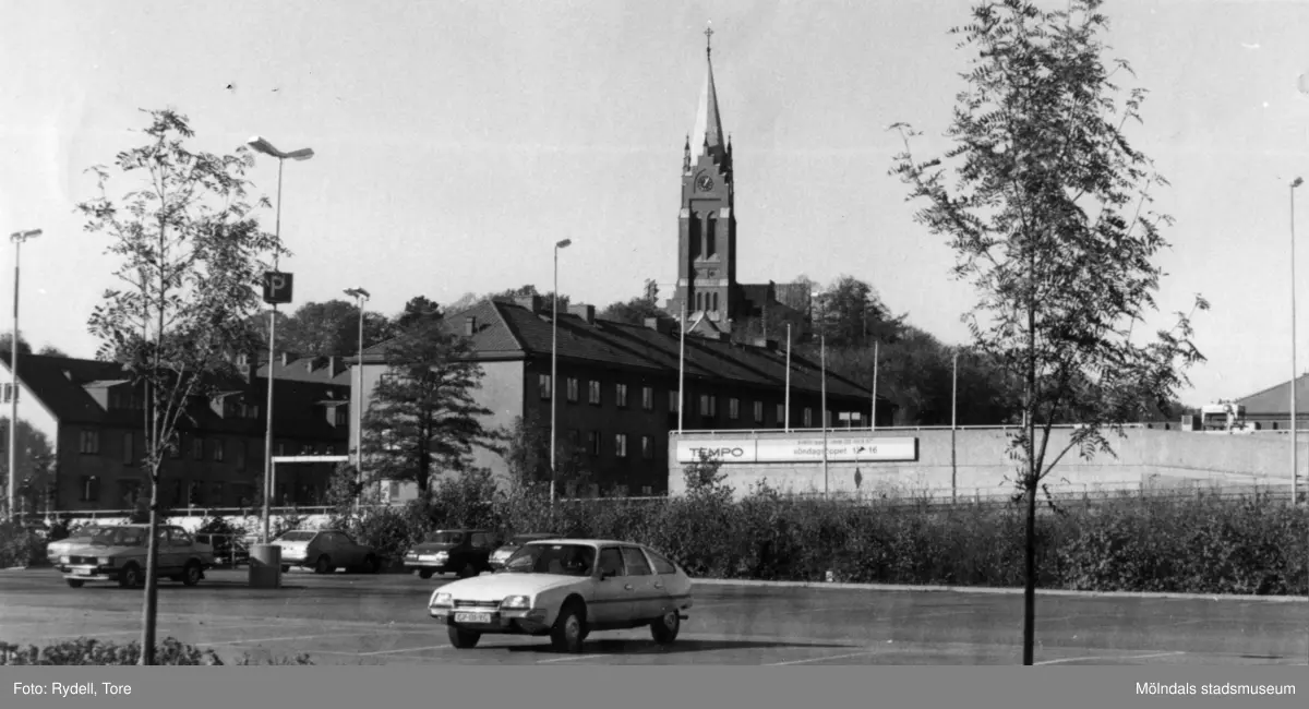 Vy från Barnhemsgatan över parkeringsplats i Mölndals centrum mot fastigheter vid Storgatan. I bakgrunden ses Fässbergs kyrka.
