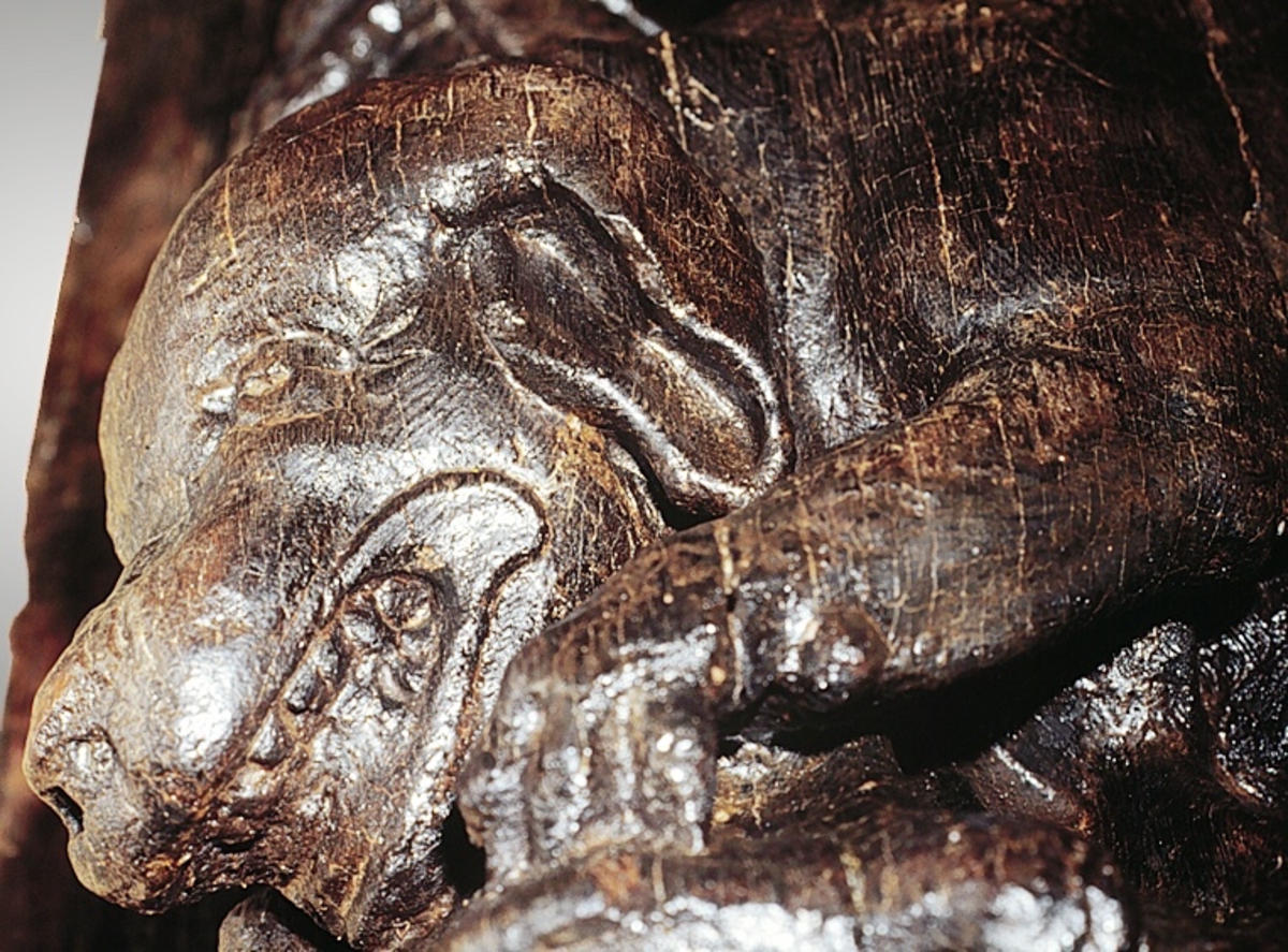 Skulptur föreställande en högrest krigare med stor, tredelad mustasch. Mannen är iklädd en romersk rustning av thoraxmodell och på huvudet sitter en slät hjälm med långa vajande plymer. Vid hans högra sida syns en veckad mantel som fladdrar i vinden. På fötterna syns en sandalliknande fotbeklädnad som sträcker sig upp på smalbenen.

Med sin vänstra hand håller krigaren en stor, oval sköld tryckt tätt mot bröstet. Skölden är prydd med symmetriskt anbragta, vågformiga räfflingar. Mitt på skölden finns en rundad upphöjning med ett hål där troligtivs en sköldprydnad, i form av en sköldbuckla eller dylikt, suttit fästad.



Hans högra arm hålls lyftad rakt ut från sidan och underarmen är böjd i en vinkel uppåt. I sin knutna hand har han hållit ett böjt svärd (eller kroksabel eller sabel) som han har dragit från svärdsskidan hängande vid hans vänstra ben. Huvudet är vridet mot vänster. Kroppstyngden vilar på höger ben medan vänster ben hålls något böjt.

Krigaren står på ett stort, frontalställt lejonhuvud. Lejonets framtassar är synliga. Framför mannens fötter, ovanpå lejonhuvudet, sitter en liten hund. Hunden är placerad diagonalt och ses i profil. Hunden har stora öron, lång, spetsig nos och lång, kraftig svans.

Figuren är skuren i ett stycke tillsammans med en stor ryggpanel vilkens baksida med urtag indikerar att skulpturen har suttit på skeppets utsida, på bordläggningen. Höger arm, manteln och svärdsskidan är dock snidade separat.

Skulpturen är mycket välbevarad. Hundens svans är bruten.



Text in English: A sculpture of a lofty warrior clad in Roman armour of the thorax type, wearing a plain helmet with long, wavy plumes. The man has a three-lobed moustache but is otherwise clean-shaven. He wears a sandal-like footwear that extend up to his shins. To the right a cloak gathered into folds flaps in the wind. In his left hand he holds a large oval shield tightly against his breast: the shield is embellished with symmetrically applied wavelike grooves and has a rounded protrusion in the middle with a hole in to which some form of a shield decoration may have been attached.

The right arm is held out from the body, at an angle, with the hand clenched, possibly having held a sword (or a scimitar or sabre) which he has withdrawn from the scabbard hanging by his left leg. The warrior has his head turned to the left, and he rests on his right leg while the left leg is somewhat bent.

The figure stands on a lion''s head facing forwards with its forepaws visible. In front of the man''s feet, and on the lion''s head, there is a small dog seen in profile, with long pointed nose, large drooping ears and a powerful tail. The figure is carved in one piece except for the right arm, the cloak and the scabbard which have been added separately. It has a large back-board of which the back reveals that the sculpture was attached to the outside planking of the ship.

The sculpture is particularly well preserved, with several traces of gold visible at the time of the salvaging. The dog''s tail is broken.