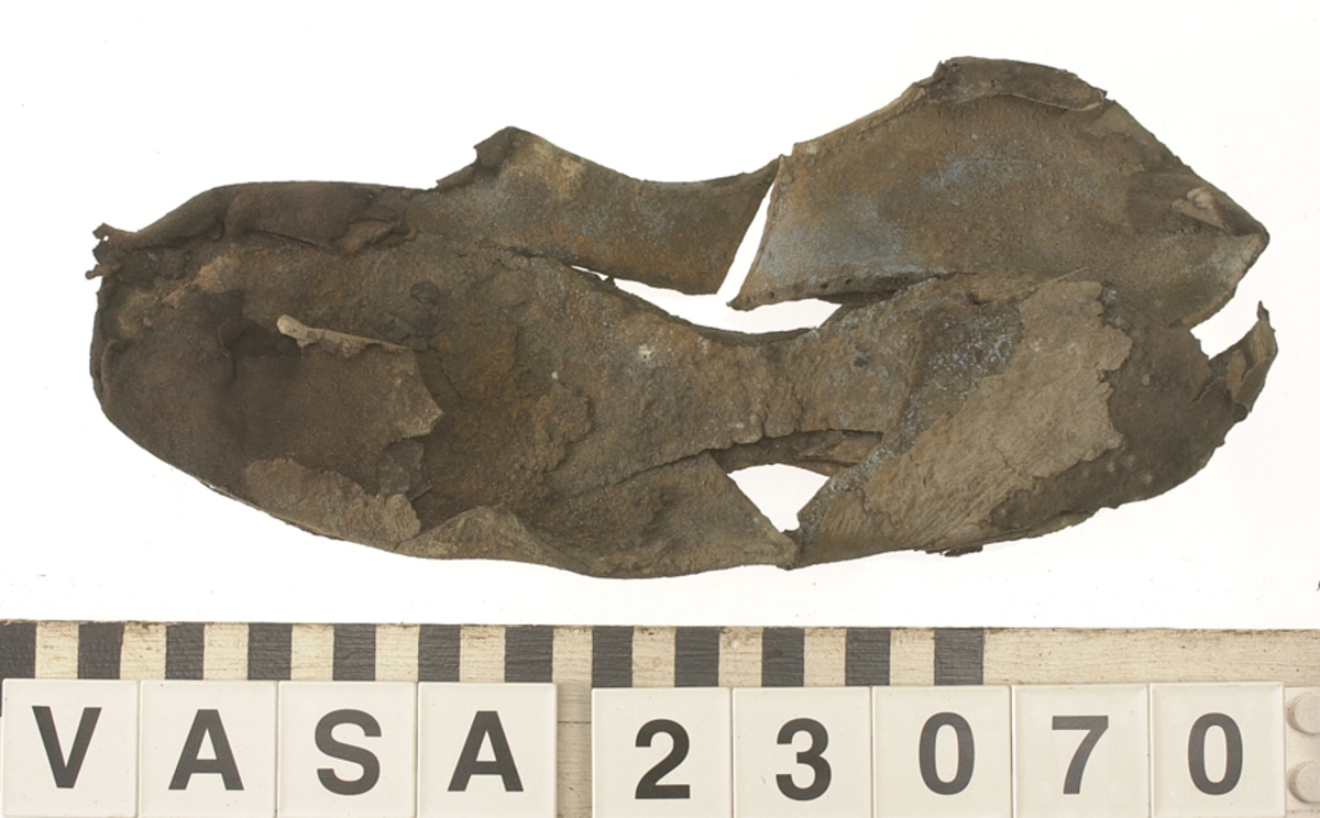 Sko.
Inuti skorna hittades även fragment från olika textilier som ursprungligen varit ett par strumpor. Idag sitter ett stickat fragment av ull fast i vänsterskon (23070) med avigsidan uppåt. I fyndet finns även en mängd lösa stickade fragment som idag är mycket korroderade och vissa helt mineraliserade av järn vilket har gjort textilen stenhård och därmed oerhört skör (23070a-c). Utifrån minskningar som några små fragment har så har det varit möjligt att rekonstruera strumpans tå. Genom att pussla ihop fragmenten som ursprungligen har suttit ihop har det på så vis gått att avgöra hur strumpans tå har varit konstruerad. Då textilen är så fragmentarisk är det inte möjligt att avgöra hur resten av strumpan har sett ut eller varit konstruerad. Däremot så finns det fragment av ett kypertvävt tyg av ull som på vissa ställen ligger dubbelt med det stickade 23070c). Ett av dessa fragment har stelnat i formen så som den suttit på fotens främre del inuti skon. Detta fragment täcker delvis det stickade tyget och har dessutom spår av kaststygn längs en kant. Så som det vävda tyget är placerat på det stickade tyget pekar på att man använt det som lagningslappar till sina stickade strumpor. 
I fyndet finns det även korroderade järnklumpar, ett tuskaftsvävt tyg av ull och rester av ett grovt bastfibergarn och två mindre läderfragment (23070d-f). Det är möjligt att även dessa har tillhört strumpan och/eller skon.

För mer detaljerad information från textildokumentationen (år 2022) se länkade filer.

Skodokumentation 2022:
Ovanläder. Tillverkad i getskinn av god kvalitet. Hör ihop med fyndnummer 23069 (Yngves skor). Tillverkades på en rak läst och bars på vänster fot. Inga dekorationer Det finns stygnhål som visar att det funnits invändiga förstärkningar (men förstärkningslappen saknas). Det finns delaminering men inga lagningar.
För mer information, se Länkade filer: Uppritning

Text in english:
This is the upper of a shoe of more “extreme fashion”, made of goatskin leather of good quality, probably made on a straight last and worn of the left foot, with no decorative features. The upper couples with 23069, together composing Yngve’s shoes. There are internal reinforcements missing from the upper and delamination, there are no repairs.
For more information, go to Länkade filer: Uppritning