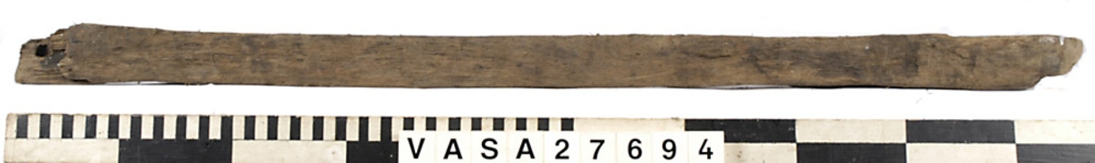 Relingslist. Den ena kortänden är uppfläkt. Den andra kortänden avslutas med en snedfasning, sannolikt en lask. Den andra slutar tvärt i sambands med en ursparning (sannolikt platsen för en årtull). Tolv stycken ej genomgående spikar i vad som sannolikt är den sida som vetter mot bordläggningen indikerar att den har spikats fast utifrån. Ett kraftigt genomgående hål syns i anslutning till ursparningarna för årtullarna. Spikhål syns i dessa avtrycks hörnor. På den sidan som vänder mot bordsidan och på undersidan syns snedfasade urtag, sannolikt för topparna till intimmer. I dessa urtagningar kan man skönja tydliga verktygsspår.