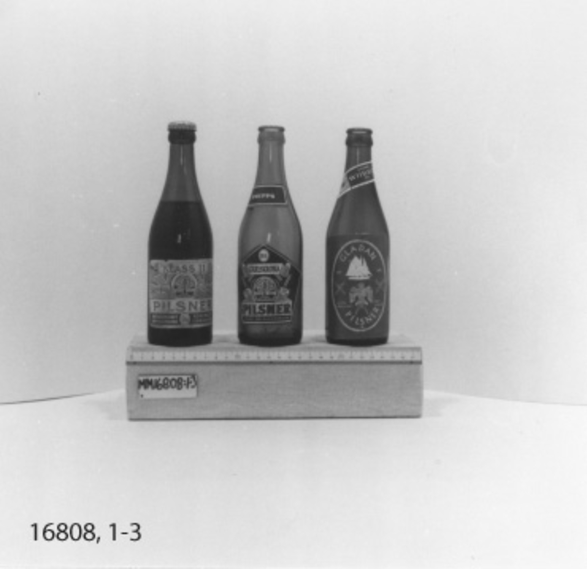 Flaska, 33 cl, i brunt glas utan kapsyl. Stor
rektangulär etikett med  vitt tryck på blå botten med text: "Gladan pilsner Glada bryggeriet  sjörullad 1947 1976" Inom ram av rep silhuett av skonerten Gladan, samt stiliserad teckning av rovfågeln gladan samt på vardera sida två korslagda gångsspelsspakar. Under denna etidett finns en oval med text= "Wibroe Pilsner Öl Helsingör". Liten etikett på flaskans hals "Dansk Wibroe öl, ölet fra Hamlets By klasse IIB". I botten  *"PLM 72 L10 SS inom vågsymbol, taggad kant.