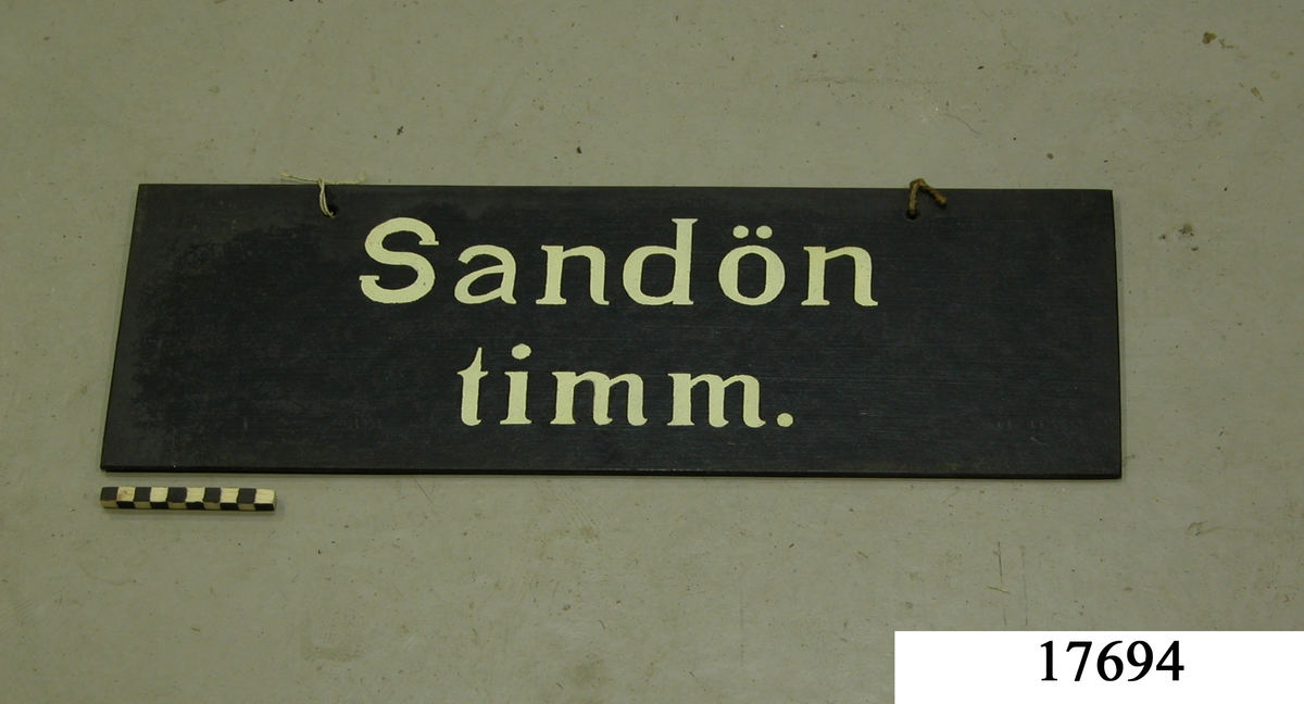 Rektangulär skylt av masonit, framsidan svartmålad, hål för upphängning. Vit schablonmålad text: " Sandön timm.".