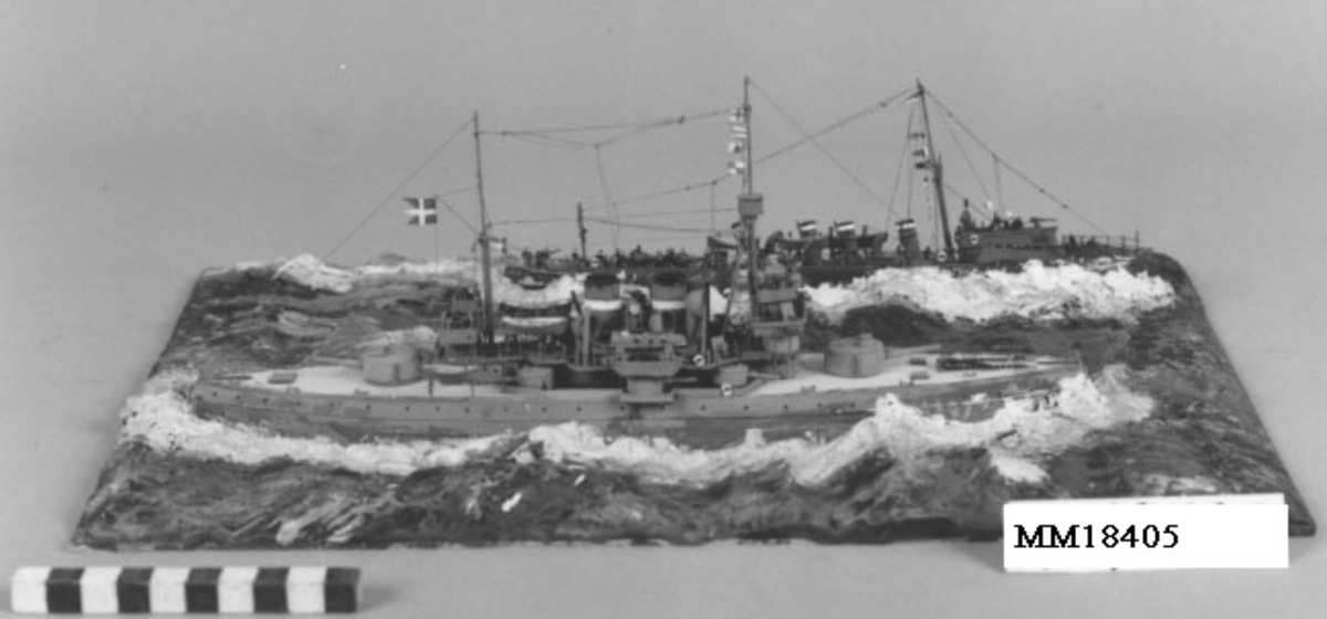 Fartygsdiorama föreställande pansarskepp av Äran-typ och jagaren Magne. Äran-typen har grått skrov, ljusbrunt däck, bryggor med mera i grått. Två skorstenar, livbåtar, artilleripjäser för och i aktern samt midskepps. Master med signalflaggor. Magne har mörkgrått skrov, fyra skorstenar, tre livbåtar, brygga i fören med mast. Små figurer på däck på båda fartygen. Fartyget är fästat till rektangulär platta och omgivet av hav av plastelina, målat i olika grönblå färgtoner. Detaljerad om än inte skalmässigt utförd. Saknar huv.