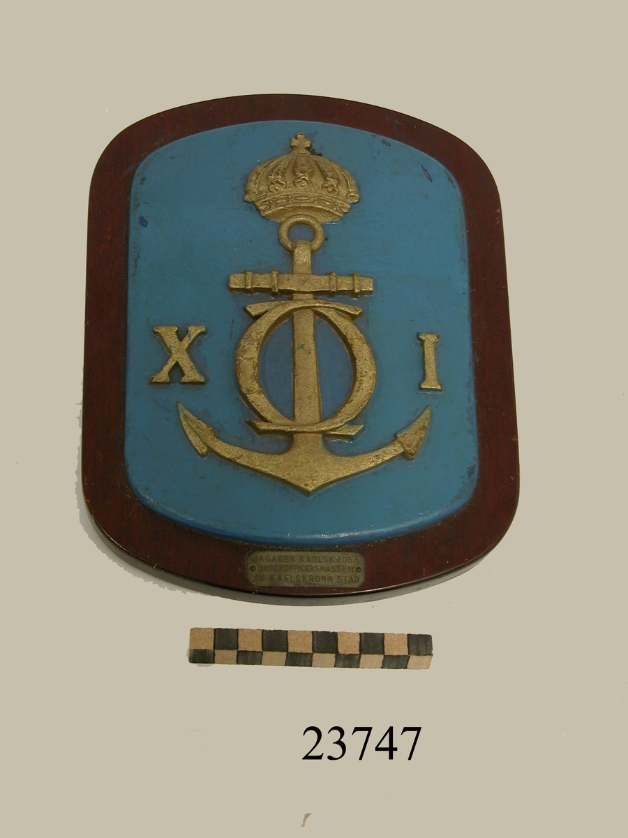 Emblem gjutet i mässing, rektangulär form med rundade hörn. Bottenfärg mellanblå. (Spår av mörkblå färg lyser igenom). Försedd med Carl den XI namnskiffer på krönt stockankare, i relief. Emblemet fäst på rekatangulär brunbetsad träplatta, rundade hörn. Nederst på träplattan liten silverbricka med ingraverad text: JAGAREN KARLSKRONA UNDEROFFICERSMÄSSEN AV KARLSKRONA STAD. På baksidan bläck för upphängning. Se SSS och X pärm.