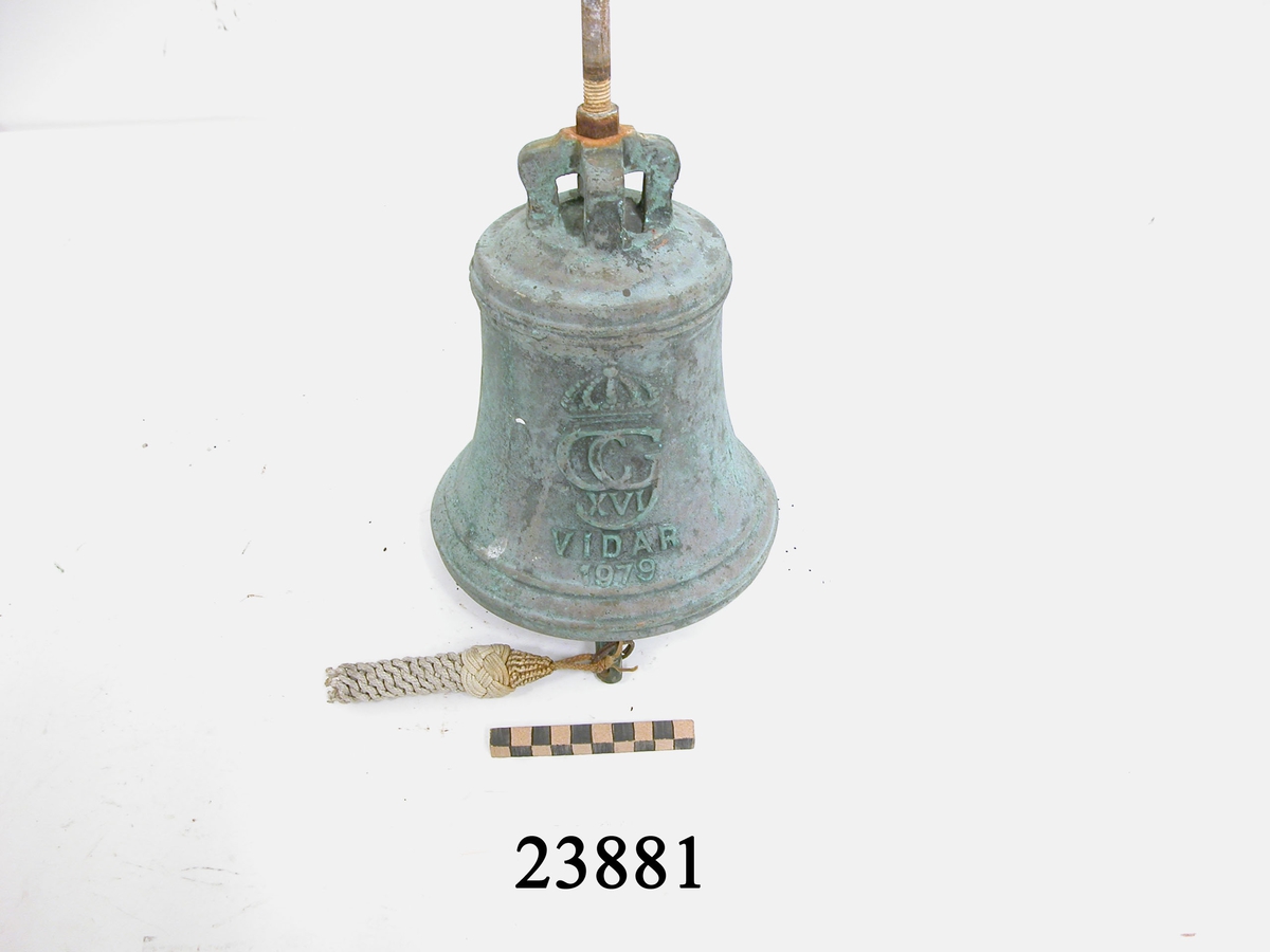 Skeppsklocka gjuten av brons, grönpatinerad. Försedd med dekor. Två linjer i dess övre kant samt två i den nedre. På klockans mitt krönt emblem: "CG XVI" därunder "VIDAR 1979". Kläpp med fockedrevsudd i makramé av vitt garn, fästad till kläppen med metallring. Överst fyra sammangående byglar för upphängning samt en 14 cm lång gängad bult med muttrar.