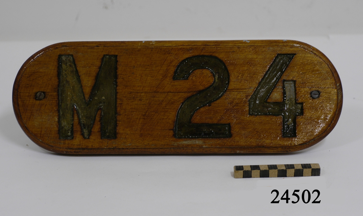 Fernissad namnbräda av trä. Rektangulär form med rundade kortsidor. I vardera sida ett genomgående hål med skruv och mutter. Kanten fasad. Bokstäverna skurna i djup relieff,målade med grågrön färg.