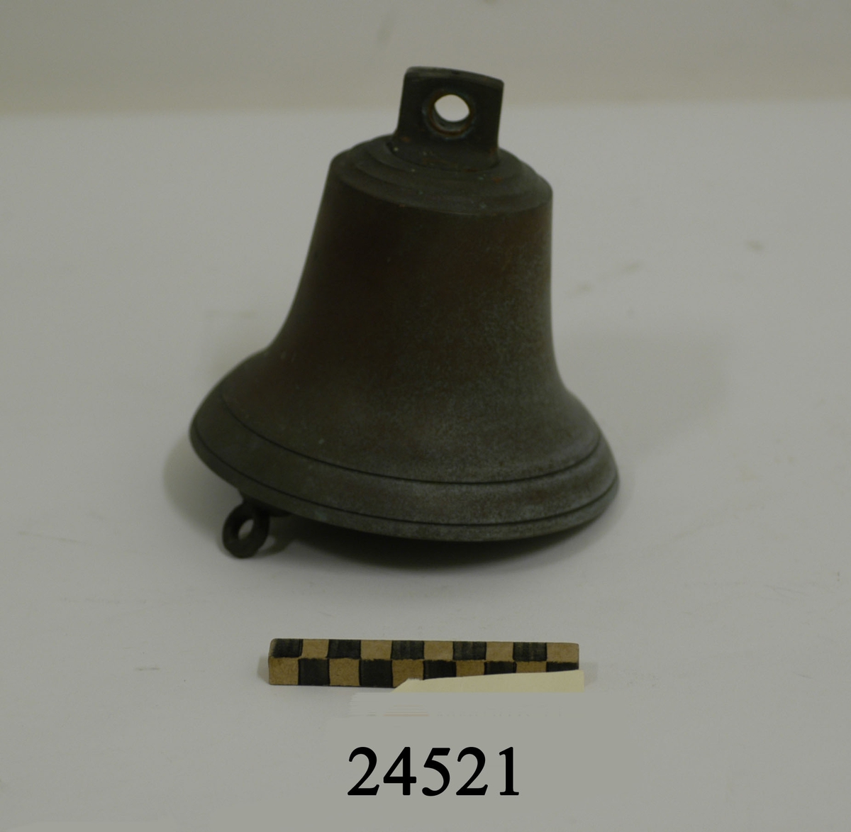 Skeppsklocka gjuten av brons. Grönpatinerad. Försedd med enkel dekor bestående av två tunna linjer på klockans nedre del. Överst en bygel för upphängning. Kläpp utan fockedrevudd. Invändigt stämpel "E.M. MORA".