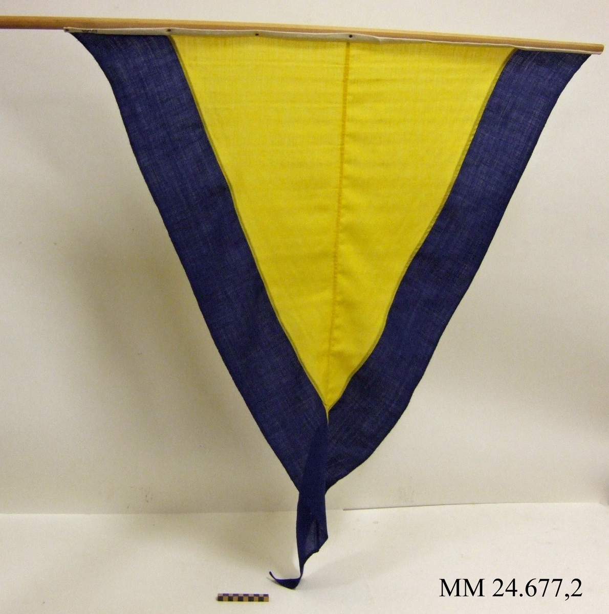 Fartstandert av ylle. Storlek 3. Även standerten Alfa. Trekantig form, gul med mörkblå kant ca 140mm. Vitt lik av bomull spikad i rundstav av obehandlat trä. Liket stämplat med tre kronor i svart. Flaggan ligger i påse tillsammans med tre andra flaggor, sk. fartflaggställ.