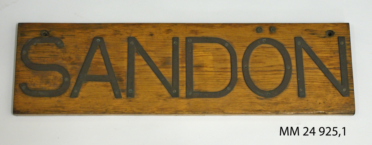 Namnbräda av fernissad treak. Bokstäver av mässing fastskruvade från framsidan, bildar namnet: "Sandön".