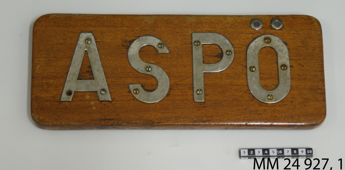 Namnbräda av fernissad teak. Bokstäver av stål fastskruvade från framsidan, bildar namnet: "Aspö".