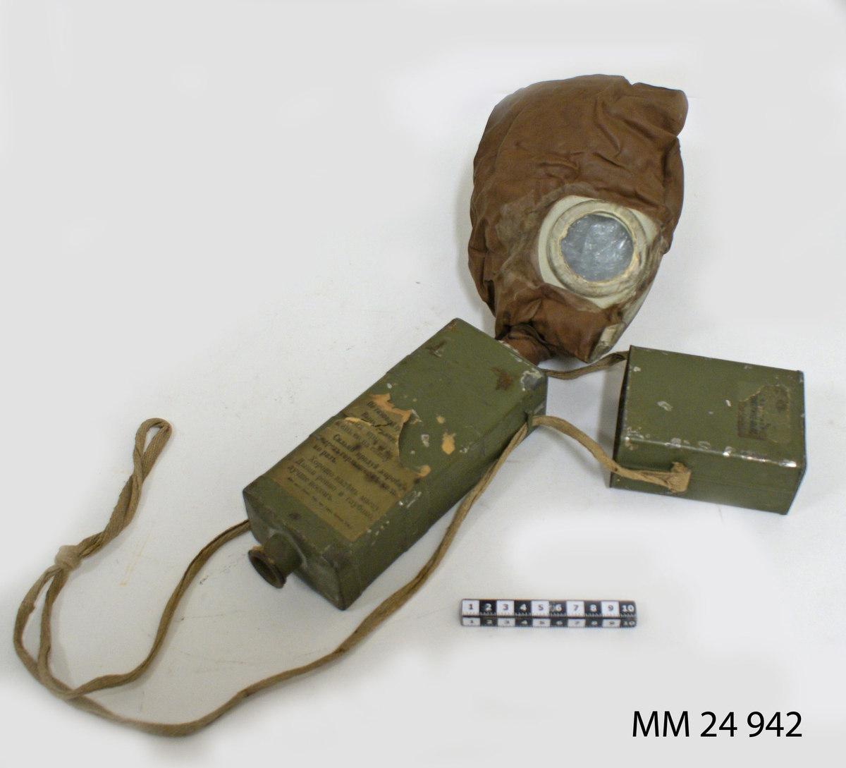 Gasmask, Zelinsky Kummant modell 16 kallad "Moscow". Rektangulär låda av plåt, grönmålad. Avtagbart lock som rymmer masken. Masken är av brunt gummi med runda titthål av glas. Gasmasken är fäst i pipa i filterlådan. Filtret är ett kolfilter med en linning av bomull som hindrar inandning av kolet. Pipa i filterlådans botten. Locket och filterlådan sitter sammen med textilsnöre som även gör att gasmasken kan hängas runt halsen. Diverse pappersettiketter med rysk text sitter på filterbehållaren: "CEPIR 296" mm.