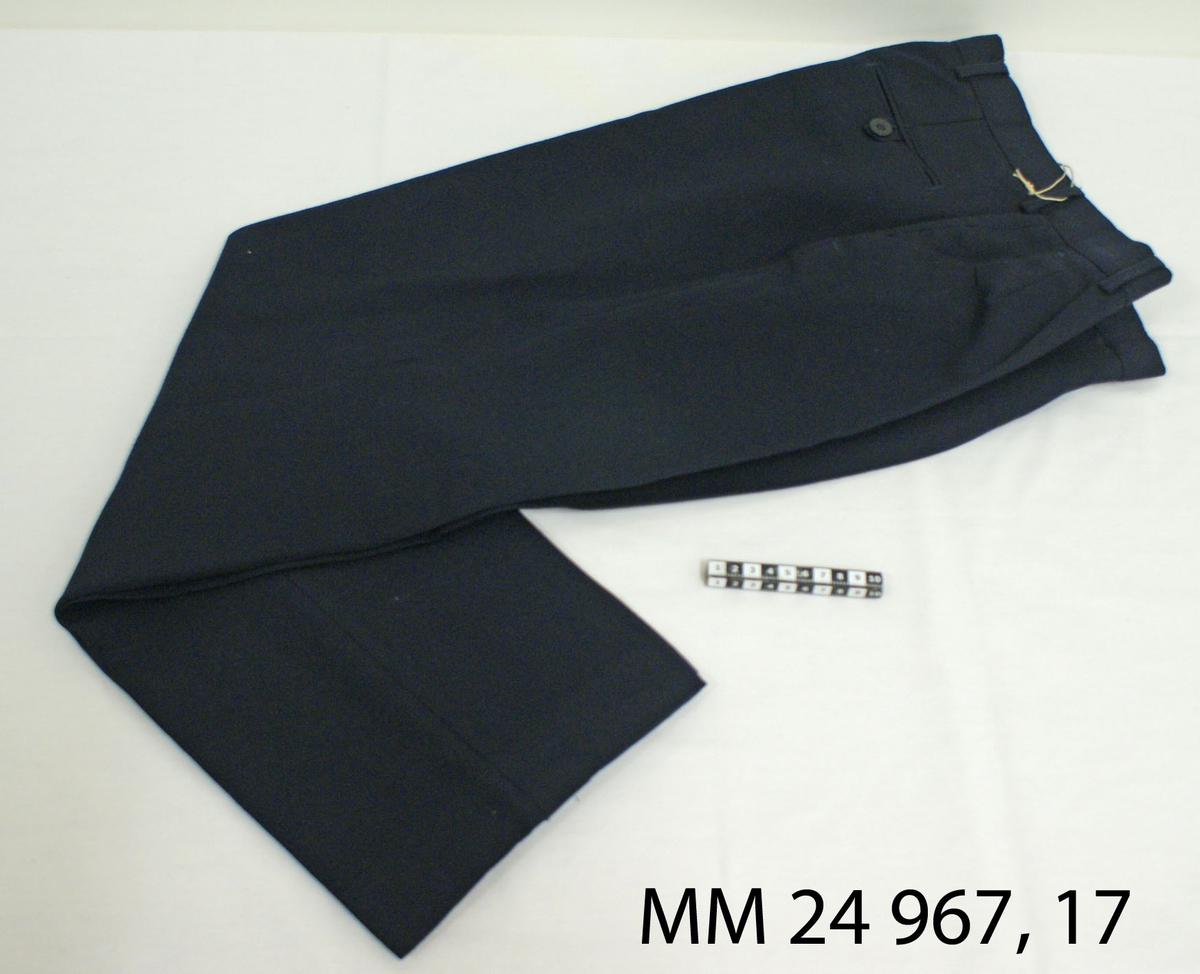 Långbyxa modell 1987, mörkblå för kvinnor. Byxor av ull och polyester med byxveck. Två snedställda sidfickor samt en bakficka som knäpps med knapp. Blixtlås i gylfen och hällor för bälte. Vit lapp fastsydd på insidan med kronstämpel, M-nummer, tvättråd samt årtalet 2002 storlek C42, "Gulins Business ab, tillverkad i Portugal".