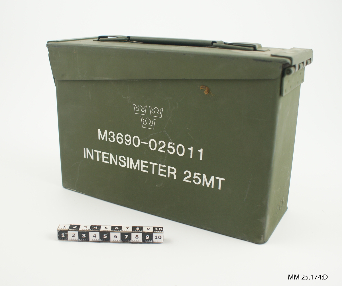 Grön låda i metall med lock. Locket fästs på ena kortsidan med hjälp av en bygel som passar in i en hake. Handtag på locket. Text på sidan: "M3690-025011 INTENSIMETER 25MT". På andra sidan sitter en skylt som anger tillverka till intensimetern samt serienummer "SN A 0349". Instansat i utstickande bokståver på baksidan: "S.C.F.". Inne i lådan sitter utskuren mjukplast där intensimetern passar i ett stort hål jämte fyra avlånga ovala hål för batterierna.