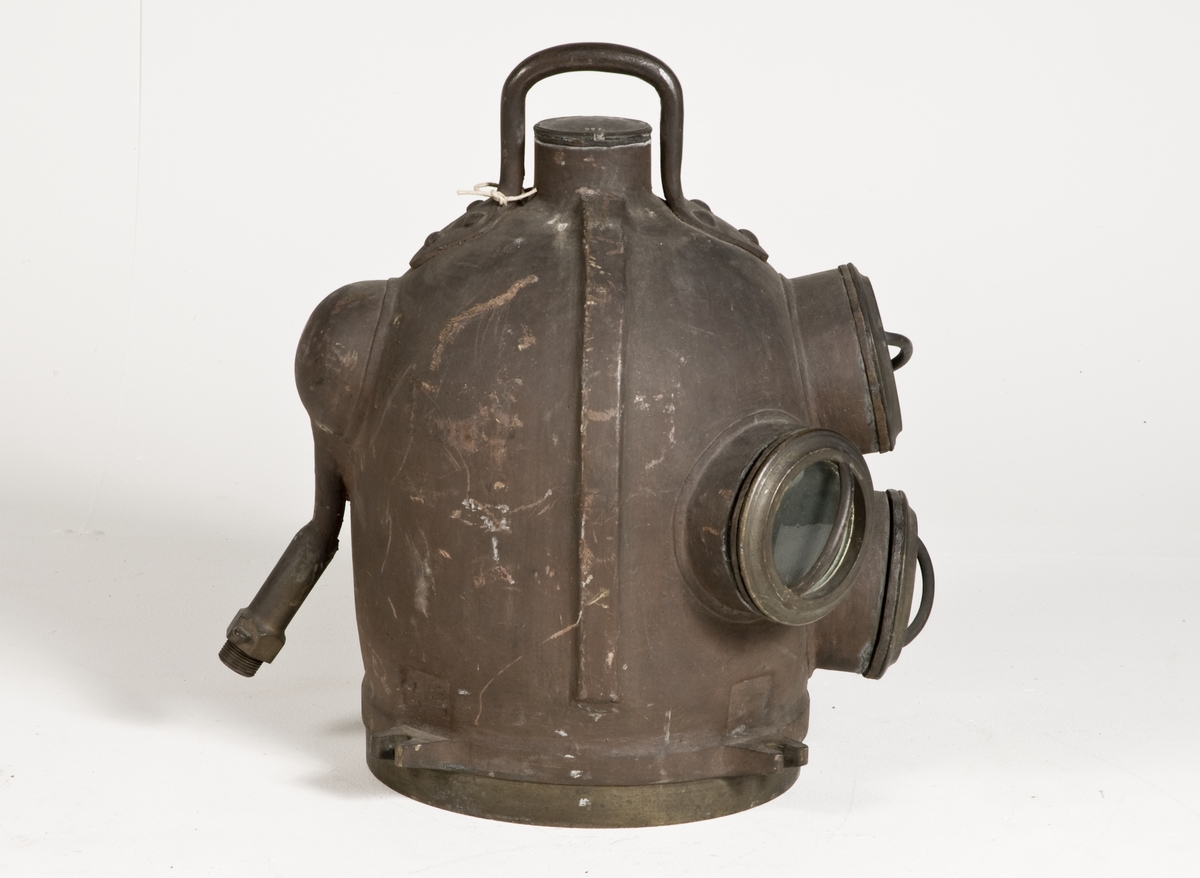 Dykarhjälm, modifierad Fahnehjelm från ca 1840-50-tal, med 4 fönster försedda med gardejärn. Upptill handtag, kort rör på baksidan för tilloppsluftslangen med backventil inuti hjälmen. Intern luftkanal från höger till vänster sida gående genom backventil med utlopp på vänstra sidan.
Från Modellkammaren.