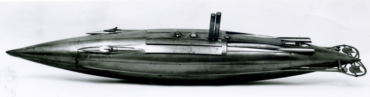 Spolformad. Stålplåt. Slingerköl, 2 propellrar av metall, 2 st höjd- och sidoroder parallellkopplade med gardjärn. 2 st höj- och sänkbara periskop av mässing. 4 st rännor för torpeder, därav 2 med luckor framtill, dels 2 för magasinering. Kort cylinder, rörlig i ränna midskepps akterut. Torpedrännorna täckas av långa stållock. Tillhörande 4 st torpeder av massiv mässing. Eneroths förslag till ubåt 1897. UndervattenstorpedFöremålets form: Spolformad.