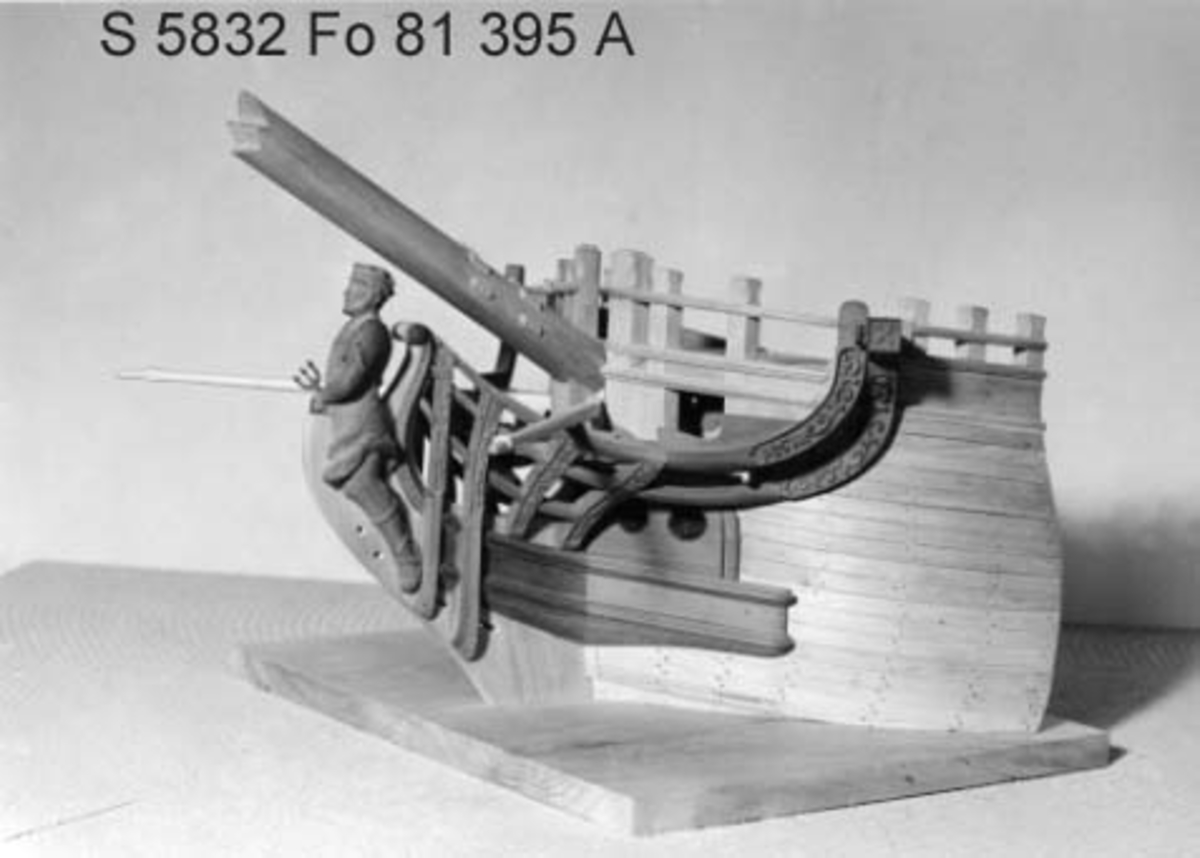 Modell av förskepp till Ostindiefarare, helt i block av al- och päronträ. Bordlagd med snidad galjon och galjonsbild föreställande Neptun, laserad i röda och bruna färgtoner. Monterad på platta av trä. Skala 1:24. Mått: Största bredd: 41 cm. Höjd till däck: 18,5 cm. Basplattan: 32,5 x 41,o cm.